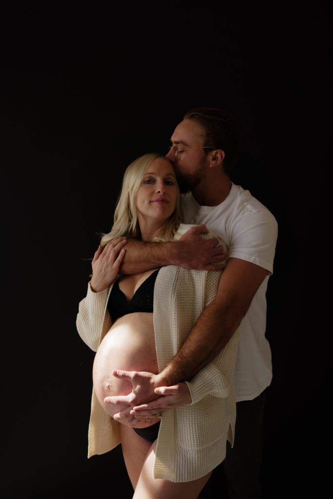 maternity photographer michigan captures an expecting couple as he wraps his arm around her shoulders from behind and holds her belly