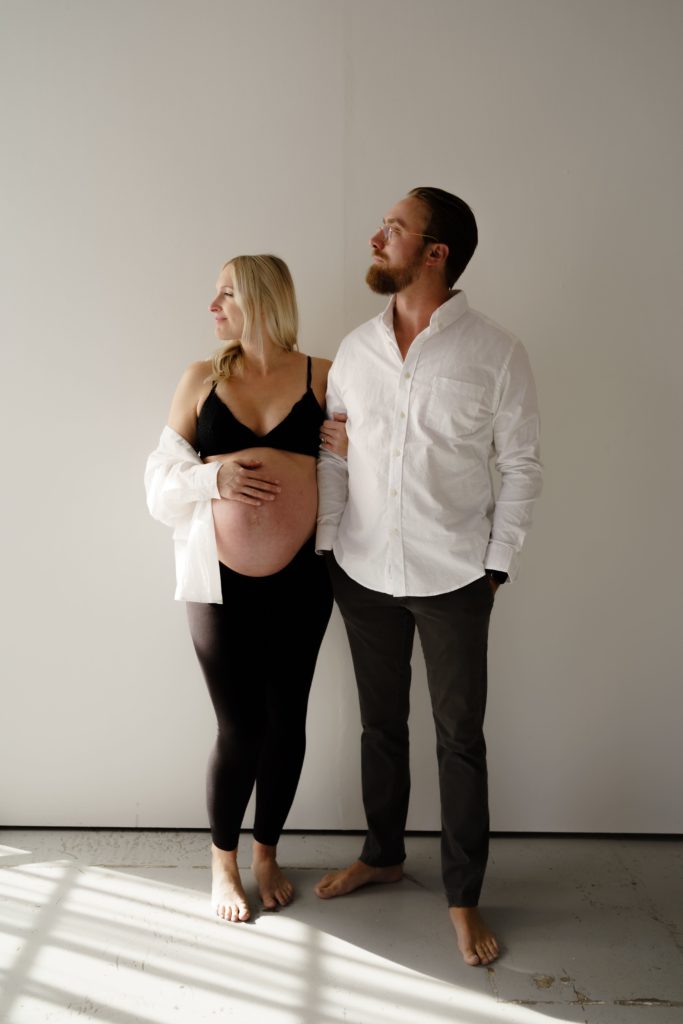 a maternity photographer michigan poses an expecting couple in front of a white backdrop, they wear white button ups and black pants and look towards the light streaming in while she holds his arm and gently touches her belly