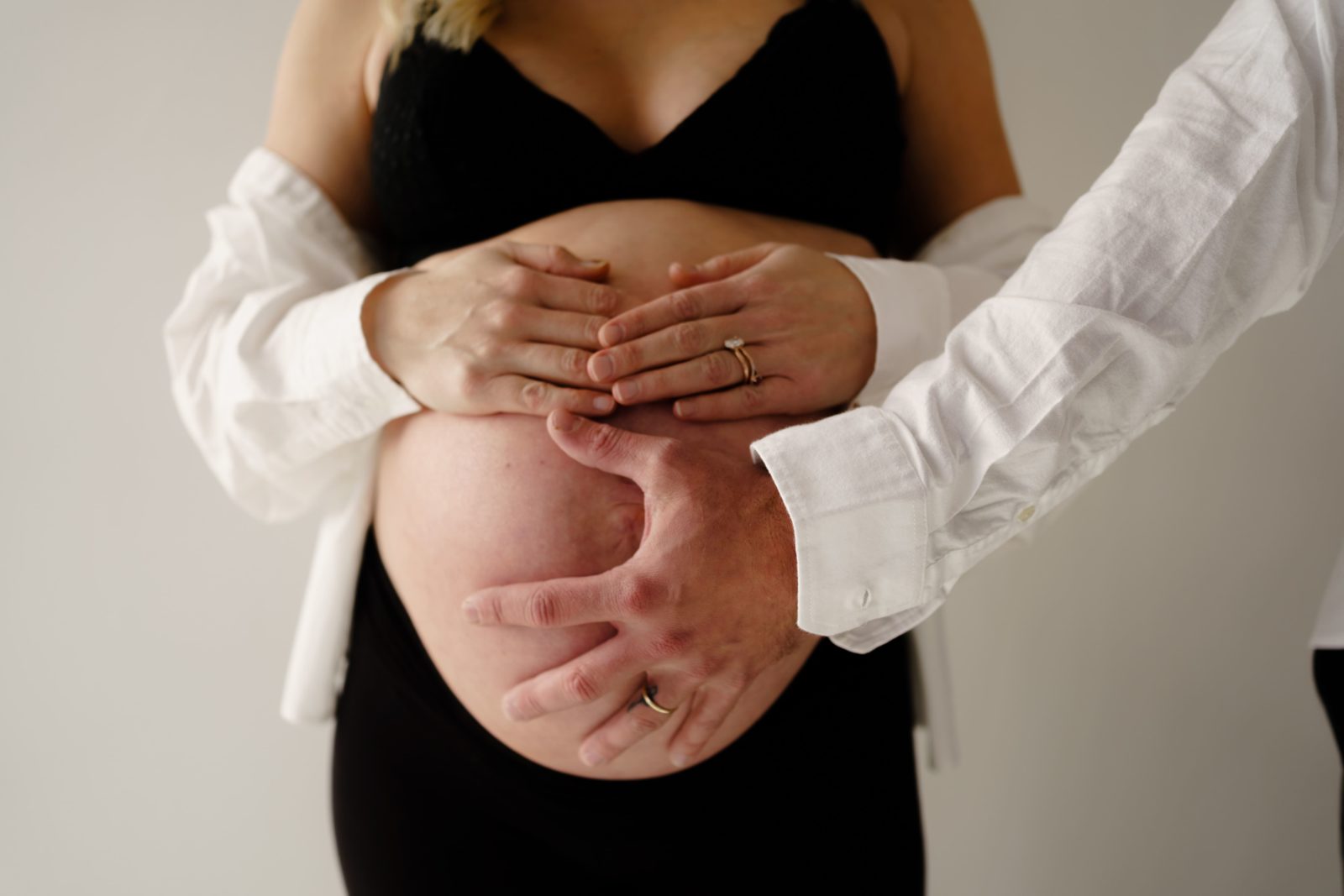during an editorial maternity shoot an expectant mother stands with an open white button down shrugged down her arms rests her hands at the top of her belly as her husband rests his spread out hand on the front of her belly