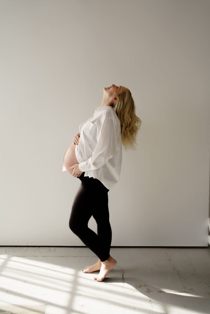 ann arbor photographer captures an expectant mother showing her belly with her hair flowing down her back as she looks up at the ceiling while standing tall