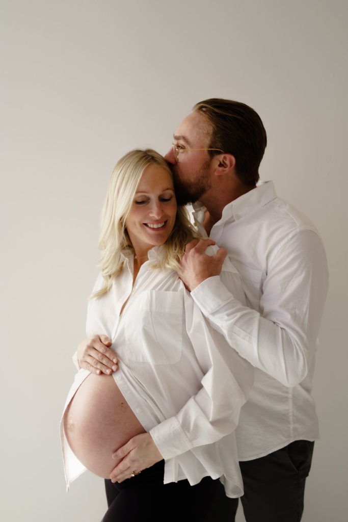 in this editorial maternity shoot an expectant couple stand wearing white button downs and black pants and he gently kisses the top of her head and holds her shoulder while she smiles holding her belly