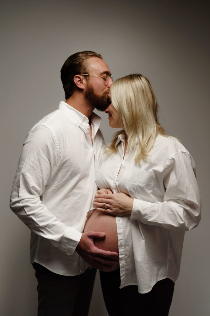 a portrait from an editorial maternity shoot in which an expectant couple stand in front of a white backdrop in dramatic lighting. he presses a kiss to her forehead as he holds her belly and she smiles while leaning into him