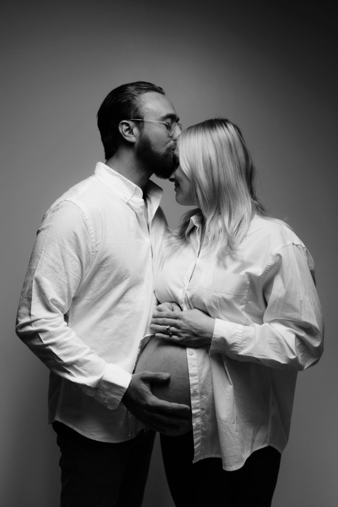 during this editorial maternity shoot the expecting couple stand in front of a white backdrop as he presses a kiss to her forehead with a hand on her belly