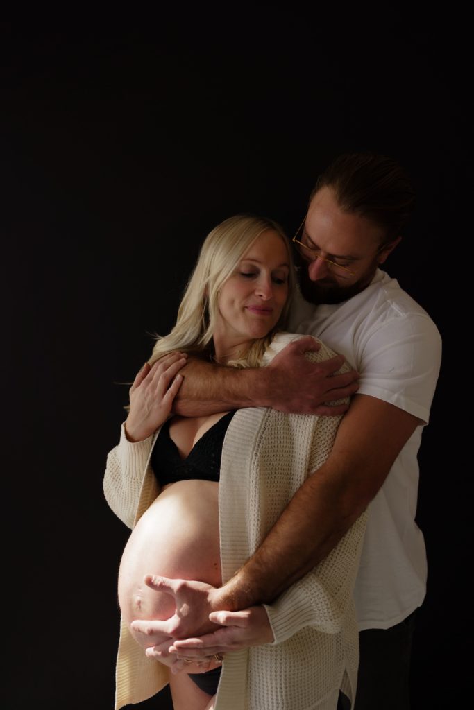 editorial pregnancy photoshoot in which an expectant couple stand in front of a black backdrop. he wraps his arm around her shoulders from behind with his other arm resting at the bottom of her belly