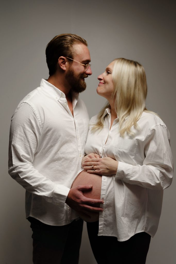 during this editorial pregnancy photoshoot an expecting couple smile at each other as he holds her belly