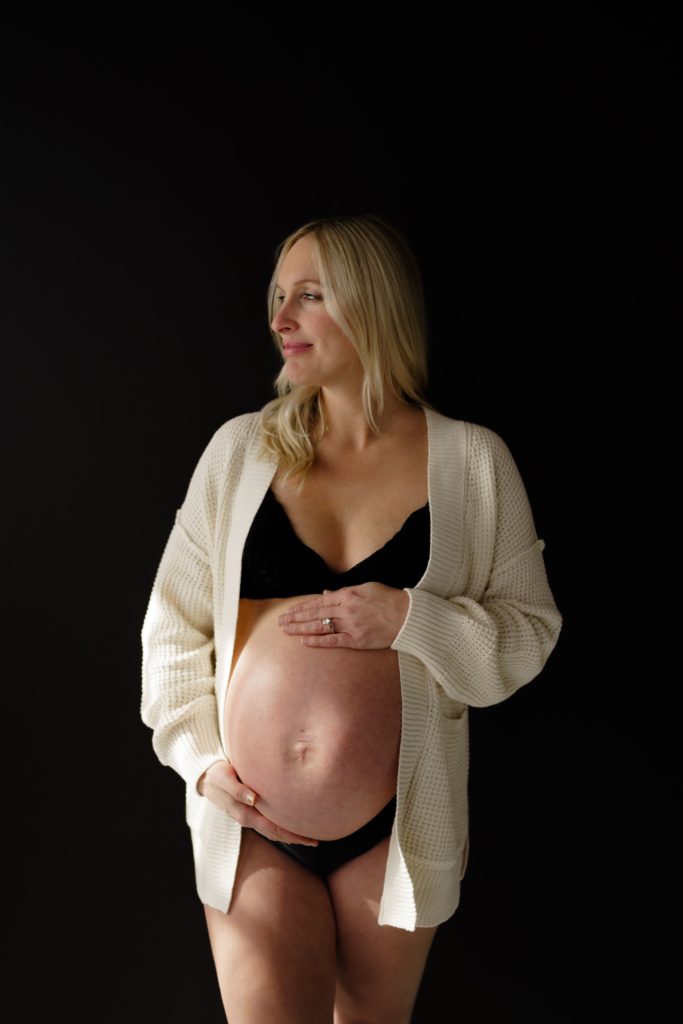 at an editorial maternity shoot, an expectant mother in front of a black backdrop while she rests her hands on her belly
