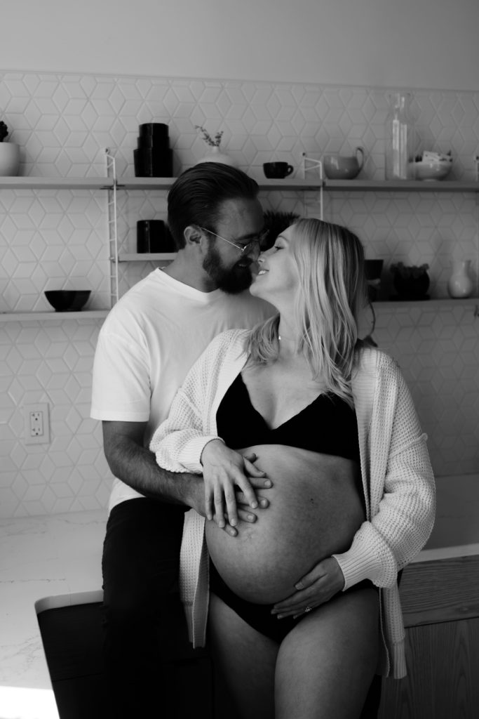 during an editorial maternity shoot a father to be sits on a kitchen counter and smiles down into his wife's face who stands leaning back into him