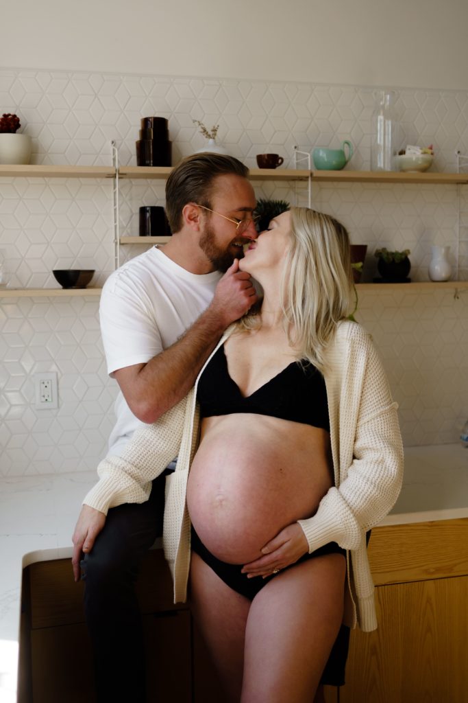maternity photographer michigan captures a portrait of an expecting couple as they lean in for a kiss in the kitchen