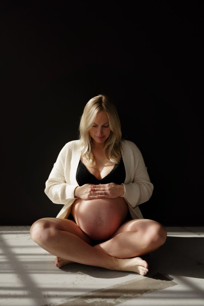 during an editorial maternity shoot a pregnant woman sits on concrete ground with a black backdrop looking down at her hands that are resting on her belly and basking in shadows and sunshine from a window
