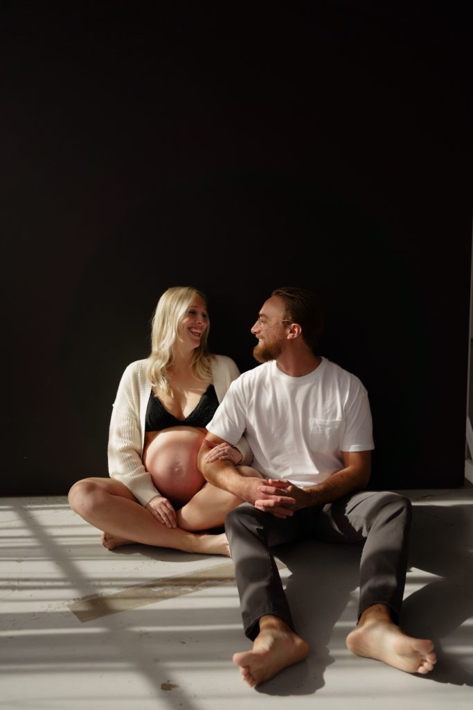 this ann arbor photographer captures an expecting couple smiling at each other during an editorial maternity shoot while sitting on concrete in front of a black backdrop, her arm wrapped in his