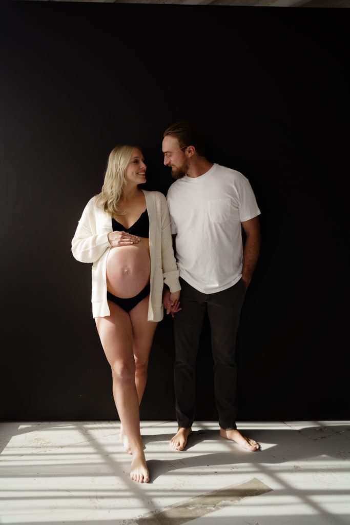 a maternity photographer michigan captures the happiness of an expecting couple as they smile at each other in front of a black backdrop, feet firmly planted on the concrete below them
