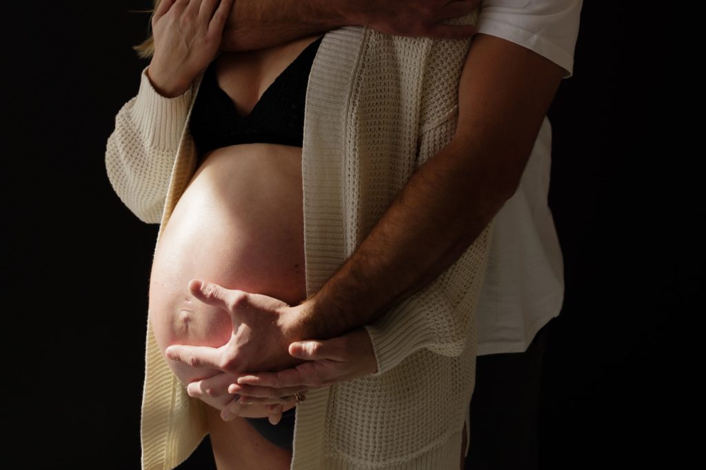 during an editorial maternity shoot an expecting couple stand leaning into each other holding the bottom of her belly