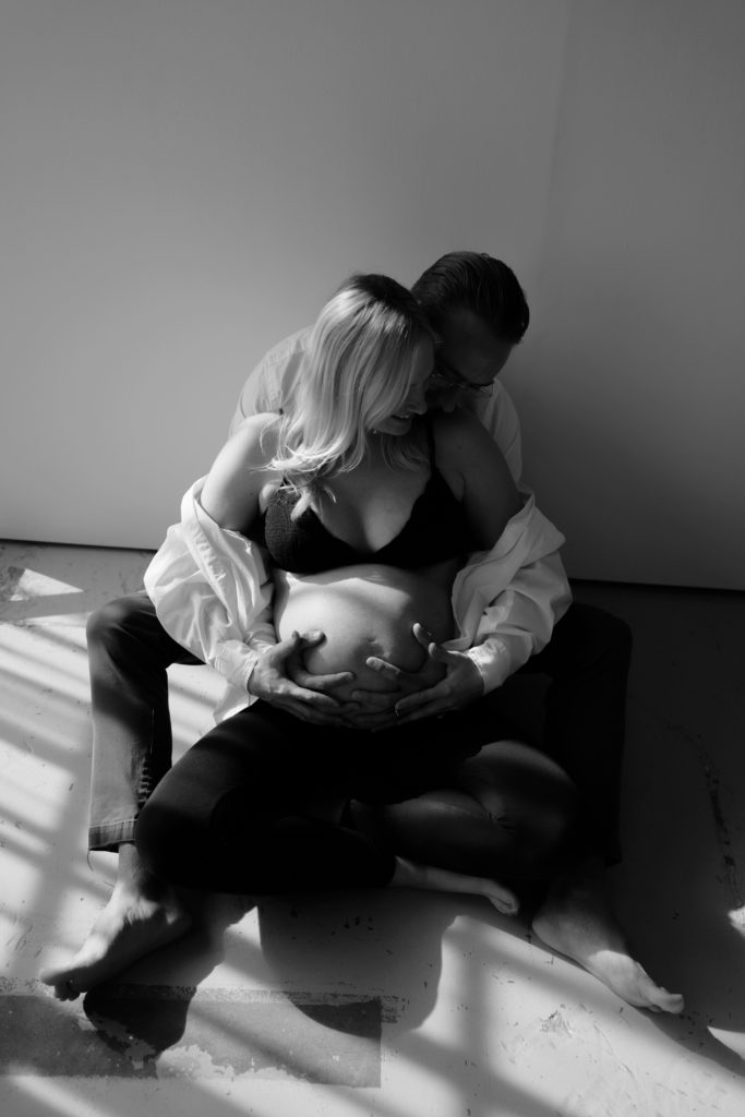 during this editorial pregnancy photoshoot the expectant couple sit leaning into each other and smiling while holding her belly
