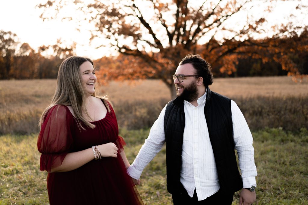 an expecting couple laugh together during their fall maternity photoshoot in front of an open field