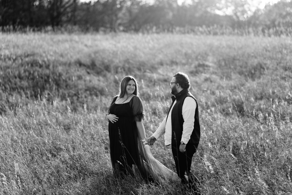 an expecting couple smile at each other while holding hands and standing in front of an open field in this black and while portrait from their luxury maternity photoshoot