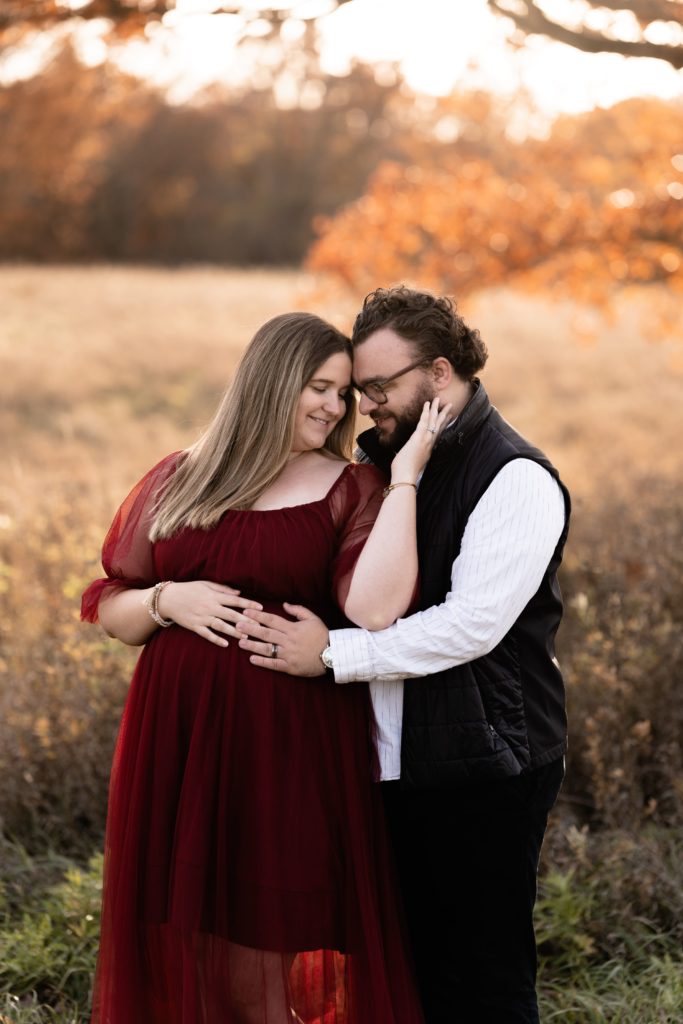 at kensington metropark michigan, the parents to be stand leaning into each other in front of an open field while resting a hand each on her belly