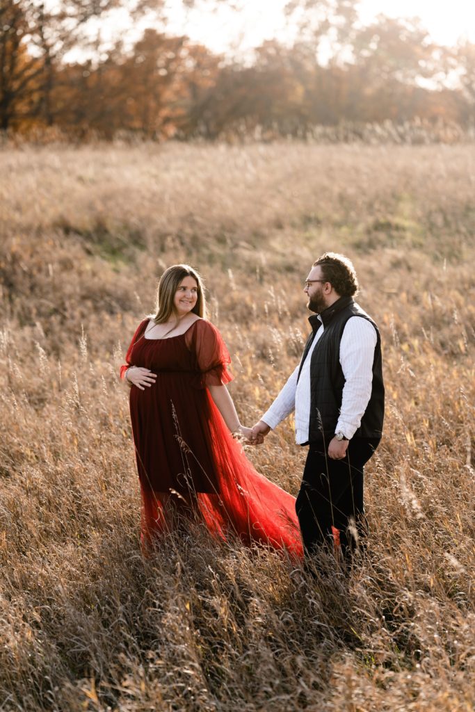 during a luxury maternity photoshoot this expecting couple smile at each other while holding hands and walking through an open field