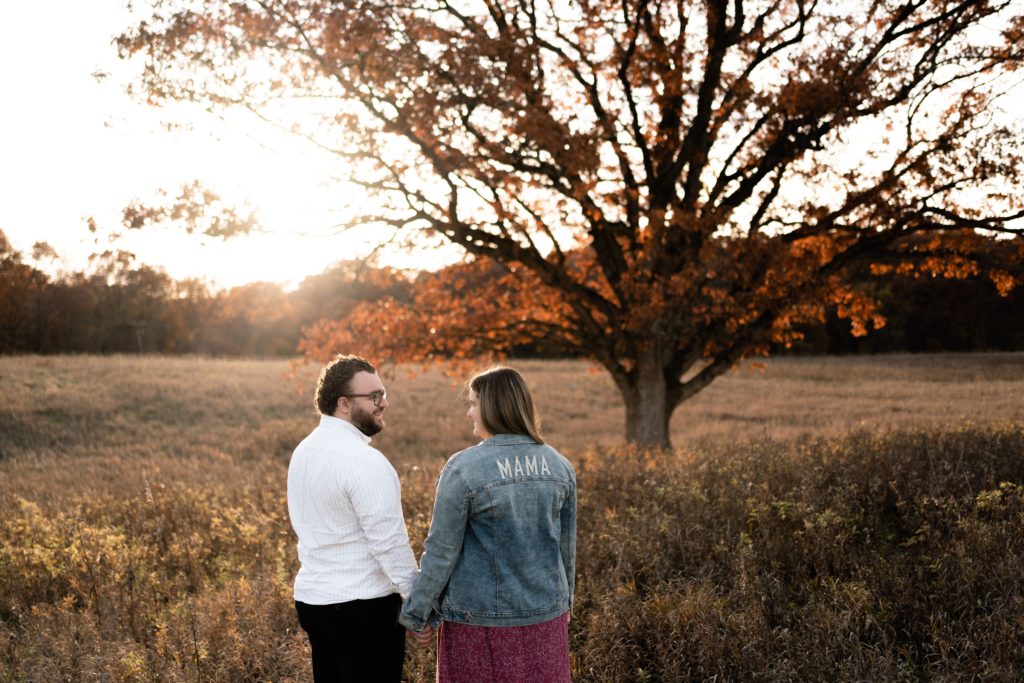 this fall maternity photoshoot captures an expecting couple standing in a field with their backs to the camera. she wears a jean jacket with the word "mama" printed on the back