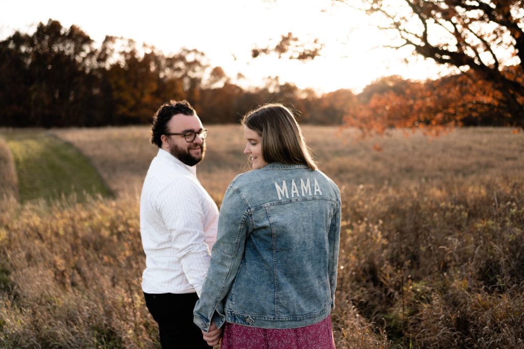 maternity photographers in michigan captures an expecting couple standing holding hands in an open field. her back is facing the camera to show off her jean jacket that has "mama" printed on the back