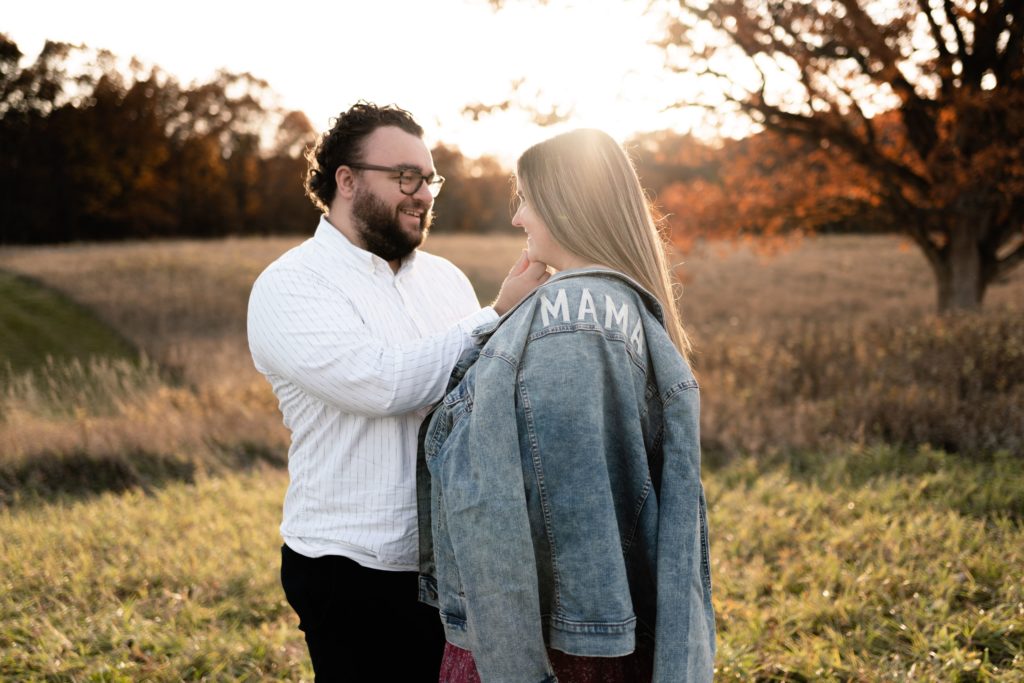 this luxury maternity photoshoot features an expecting couple standing in a fall field. he gently holds her chin as they smile at each other with her jean jacket that reads "mama" rests on her shoulder