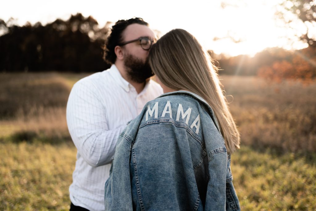 during their fall maternity photoshoot in metro detroit, the expecting father kisses his wife's forehead as her jean jacket that reads "mama" rests on her shoulder