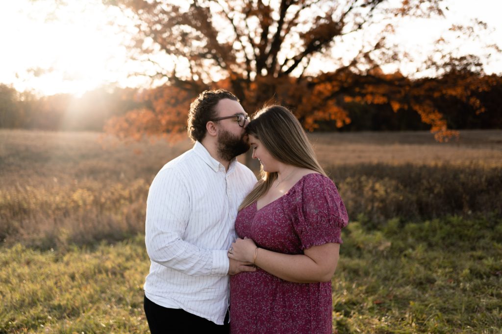 during their luxury maternity photoshoot, this couple stands in an open field. he presses a kiss to her forehead as they rest their hands on her belly