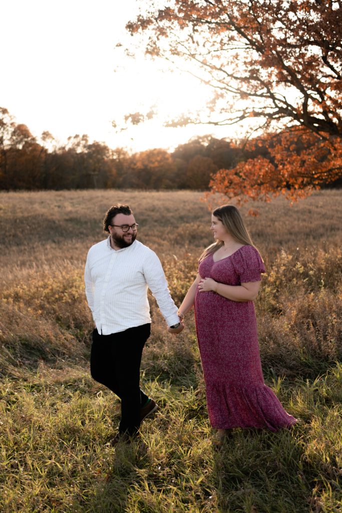 at this fall maternity photoshoot the expecting couple stand in front of a rolling field as they hold hands and walk together while smiling