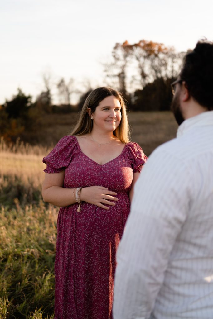 this luxury maternity photoshoot features an expectant couple smiling at each other, here she is in focus and resting her hand on her belly as she looks at her husband that stands in the foreground
