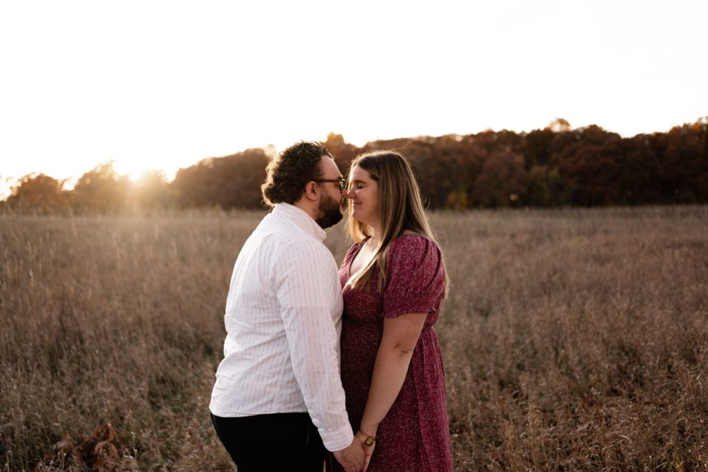 an expecting couple stand in an open field at kensington metropark michigan, their noses touching as the sun sets behind them