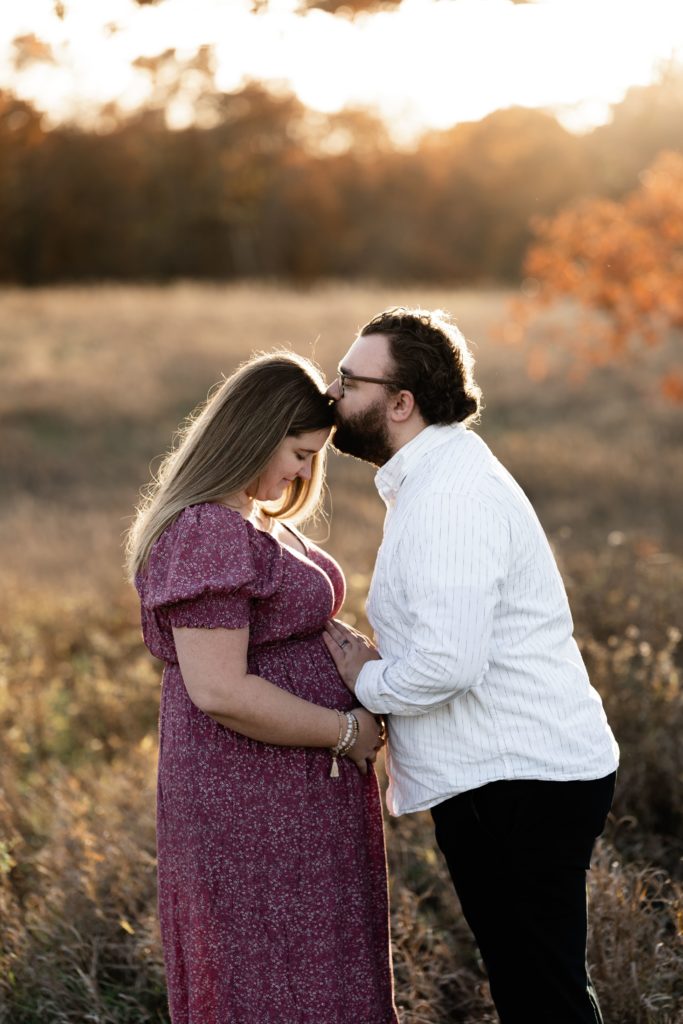 an expectant couple stand in an open field with their hands on her belly during their luxury maternity photoshoot as he presses a kiss to her forehead