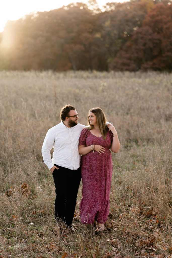during this luxury maternity photoshoot an expecting couple stand in an open field with golden light streaming behind them. he rests his arm around her shoulder as they smile at each other, her hand resting on her belly