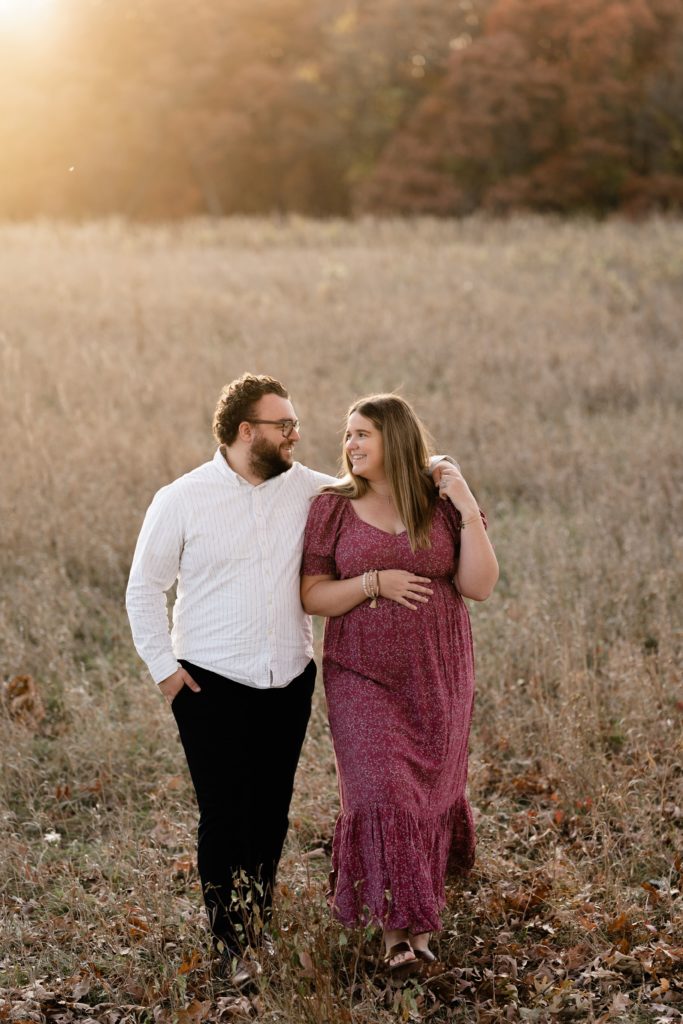 during this fall maternity photoshoot an expecting couple stand in an open field with golden light streaming behind them. he rests his arm around her shoulder as they smile at each other, her hand resting on her belly