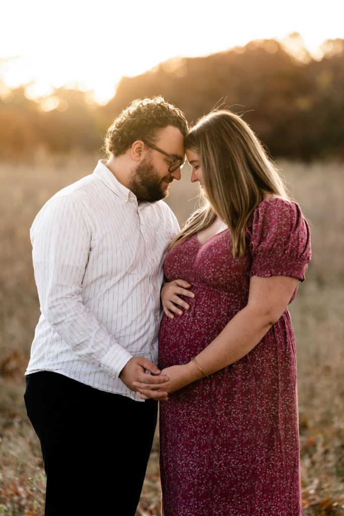 golden light streams behind an expecting couple during their fall maternity photoshoot as they press their foreheads together, holding hands in front of them as she rests her free hand on her belly