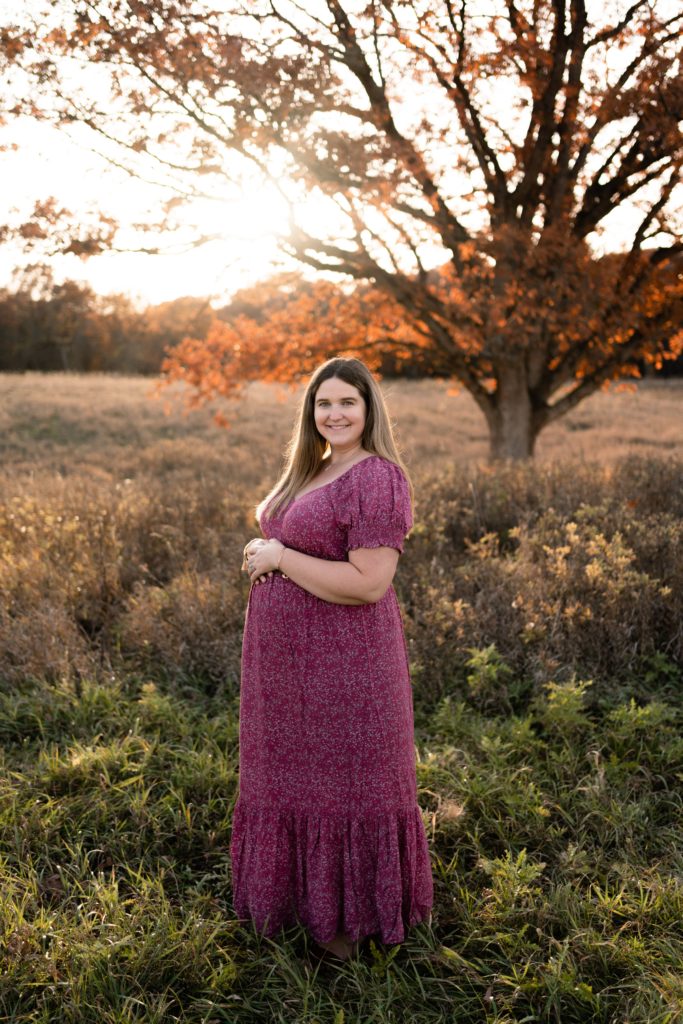 an expecting mother smiles while wearing a long, flowing purple dress and resting her arms at her belly during her fall maternity photoshoot