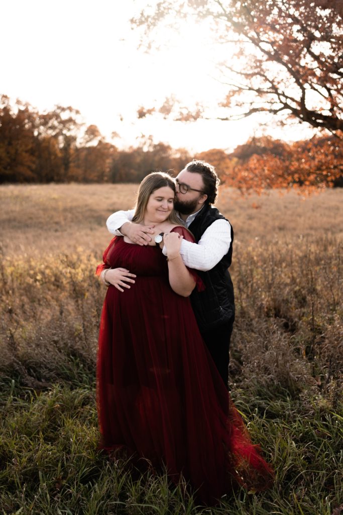 this fall maternity photoshoot features an expectant couple standing in front of an open field. he stands behind her with his arms wrapped around her shoulders. she smiles back at him as he presses a kiss to her temple