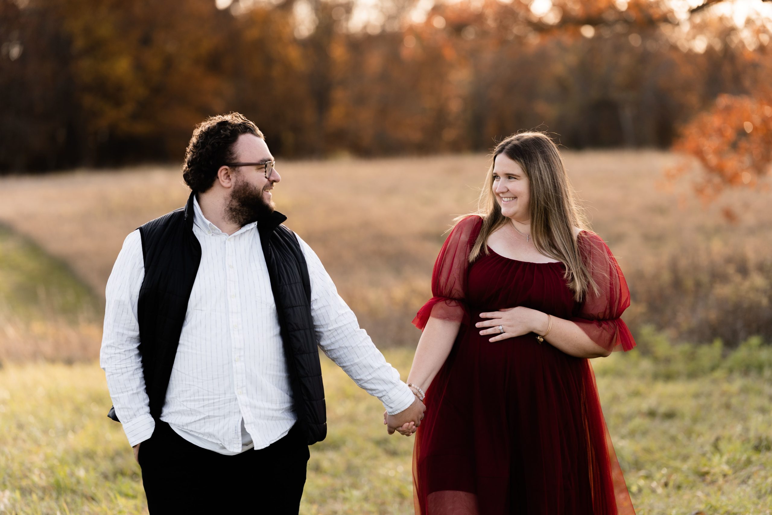 an expecting couple smile at each other while holding hands in front of an open field at their fall maternity photoshoot