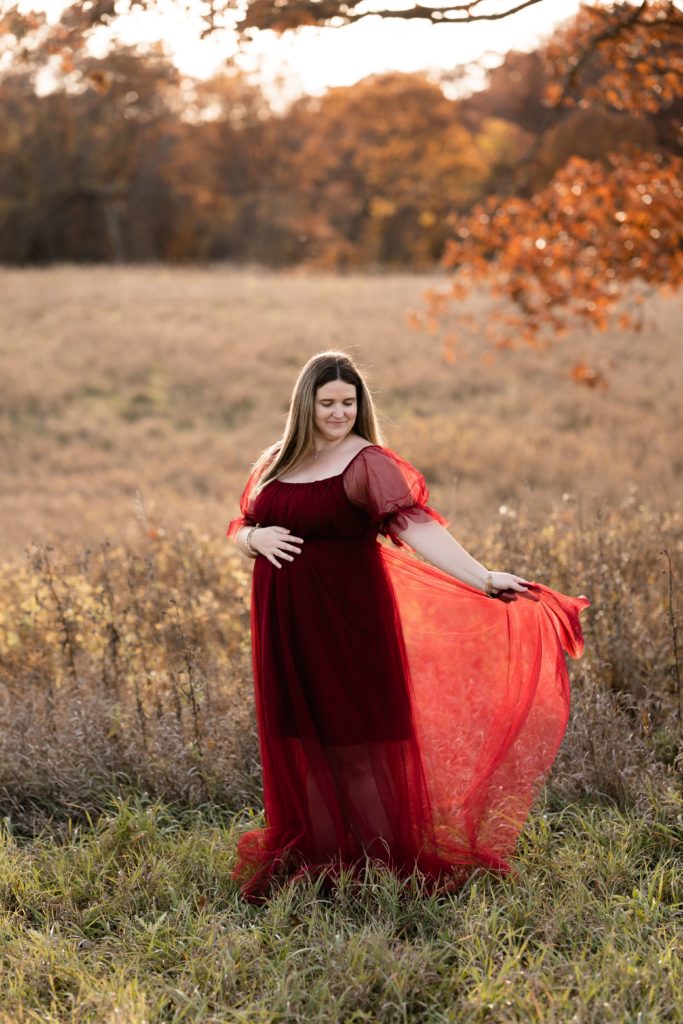 an expecting mother stands in front of an open field during a fall maternity photoshoot she smiles while looking down, touching the top of her belly and waving the skirt of her flowing dress