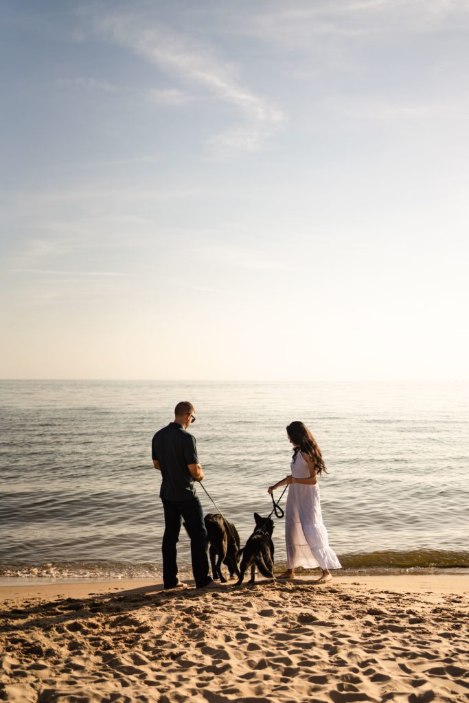 these engagement photos with dogs feature the couple each holding a pup's leash while walking along the shore
