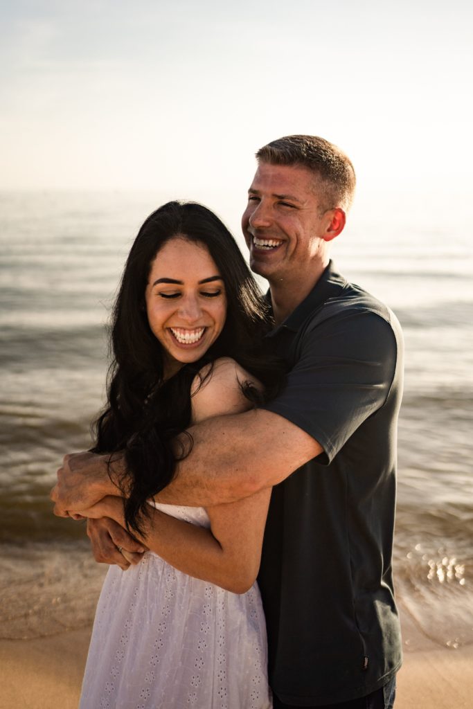 his arms wrap around her shoulders from behind as they smile wide during their engagement photoshoot beach side