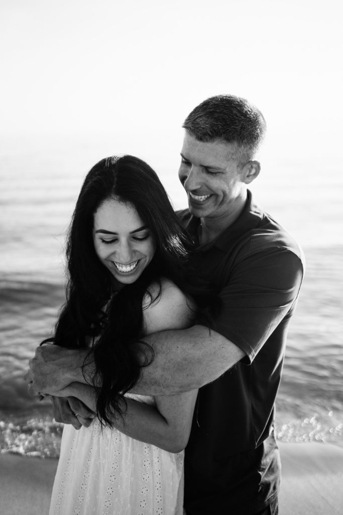 a black and white portrait of an engaged couple smiling in an embrace at the beach while michigan engagement photographer snaps their photos