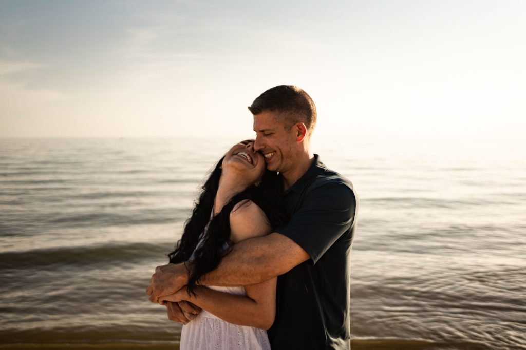 an engaged couple stand in a smiling embrace in front of the calm waves of grand haven beach for their michigan engagement photos