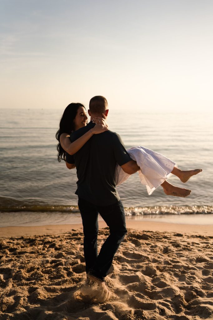 an engaged man carries his fiancee on the shore with grand haven photographers behind them
