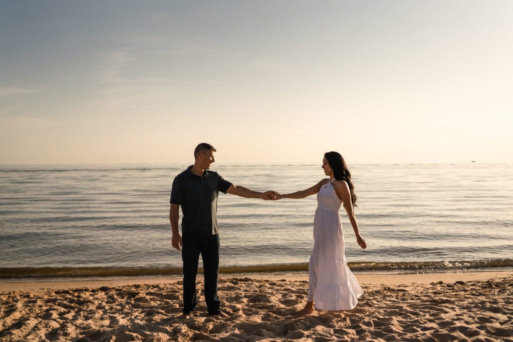 an engaged couple stand during their engagement photoshoot beach side with their hands clasped and arms at length as they dance