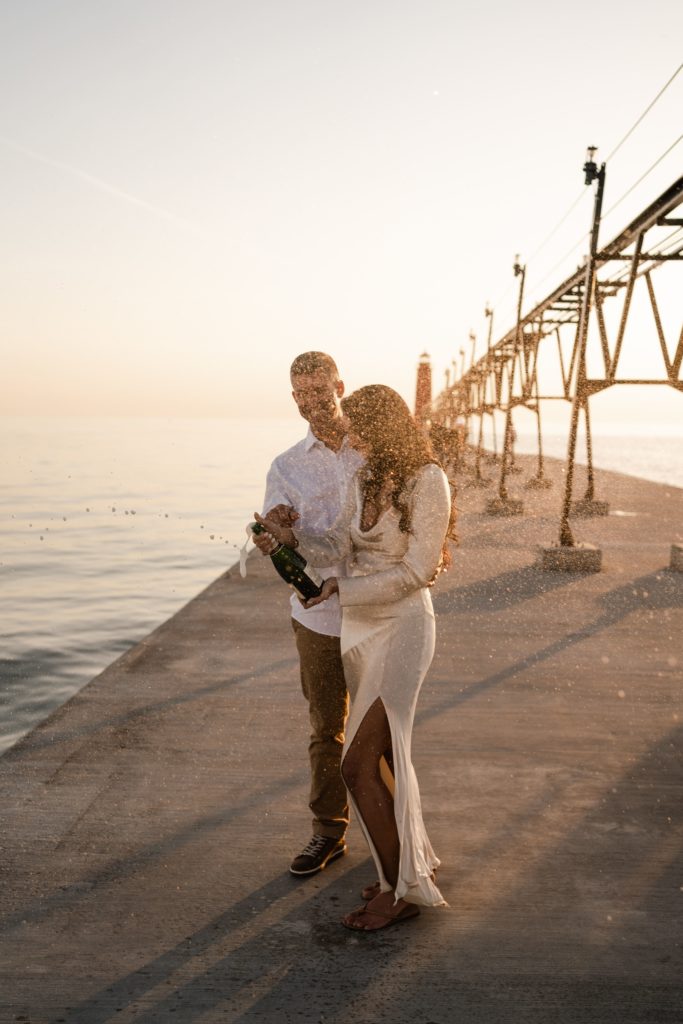 michigan engagement photographer captures a couple mid champagne pop as they stand on the grand haven pier