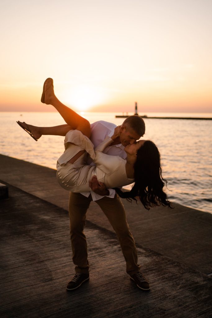 engaged couple share a kiss at the grand haven pier as he holds her in the air during their michigan engagement photos