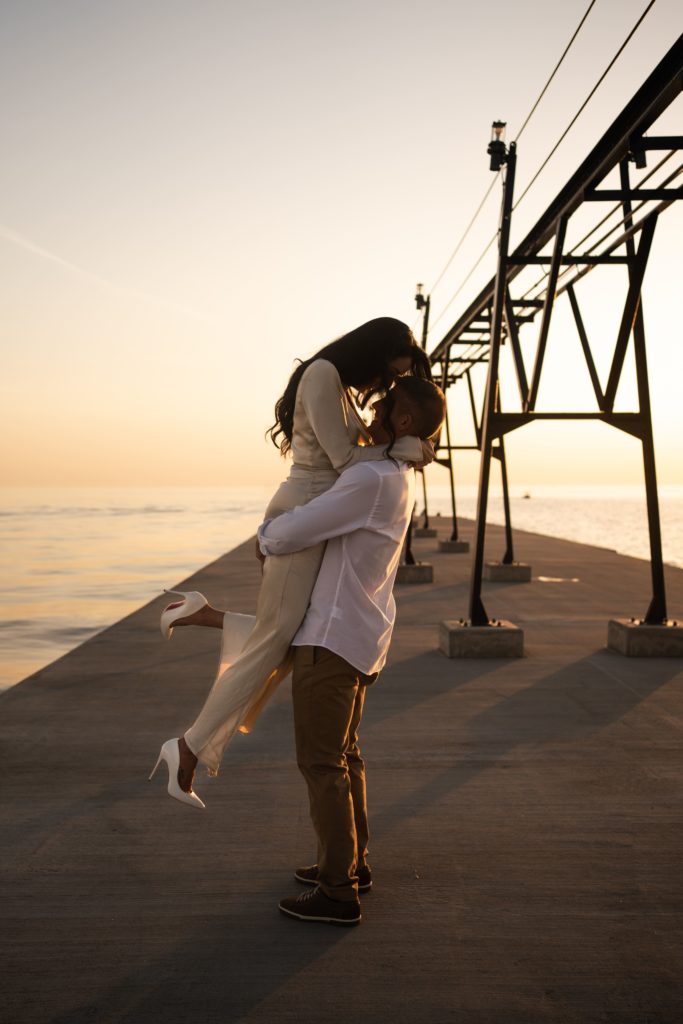 a man lifts his fiancee as she kicks out her foot during golden hour at their engagement photoshoot beach side