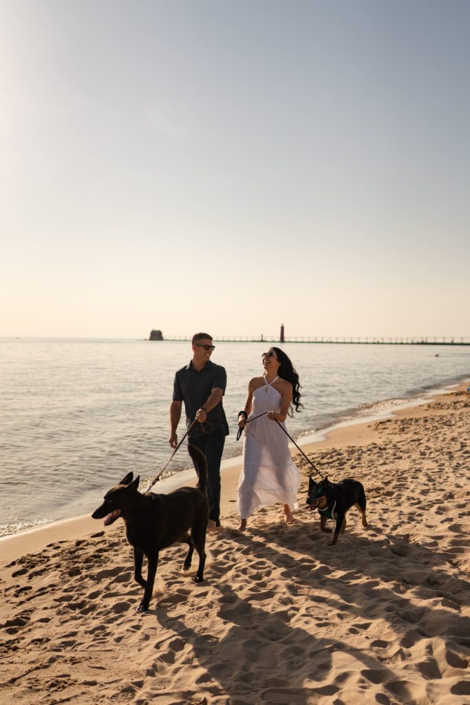 an engaged couple laugh as they walk along the shore with their pups for engagement photos with dogs