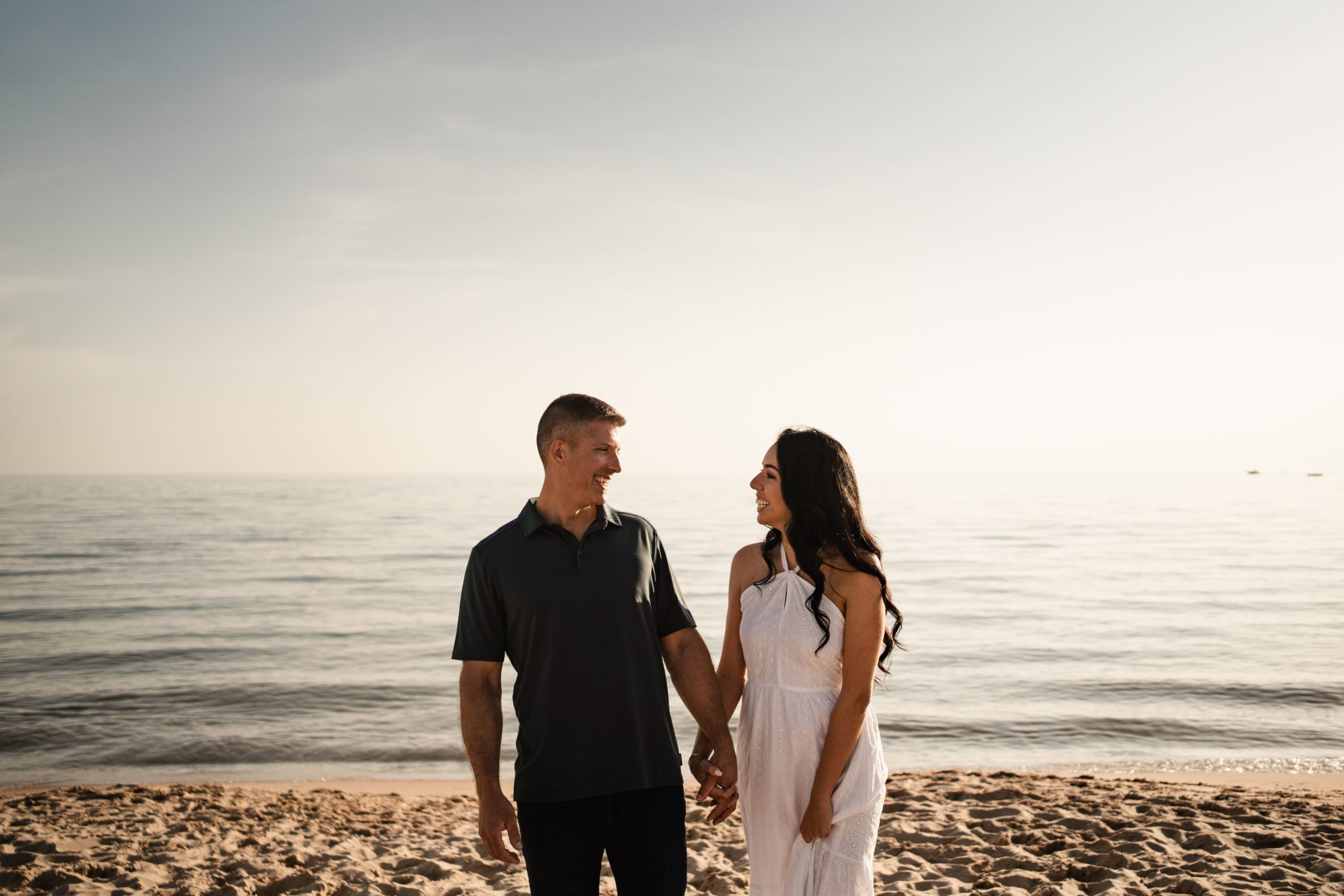 an engaged couple beam at each other as they stand holding hands in front of the beach for their michigan engagement photos