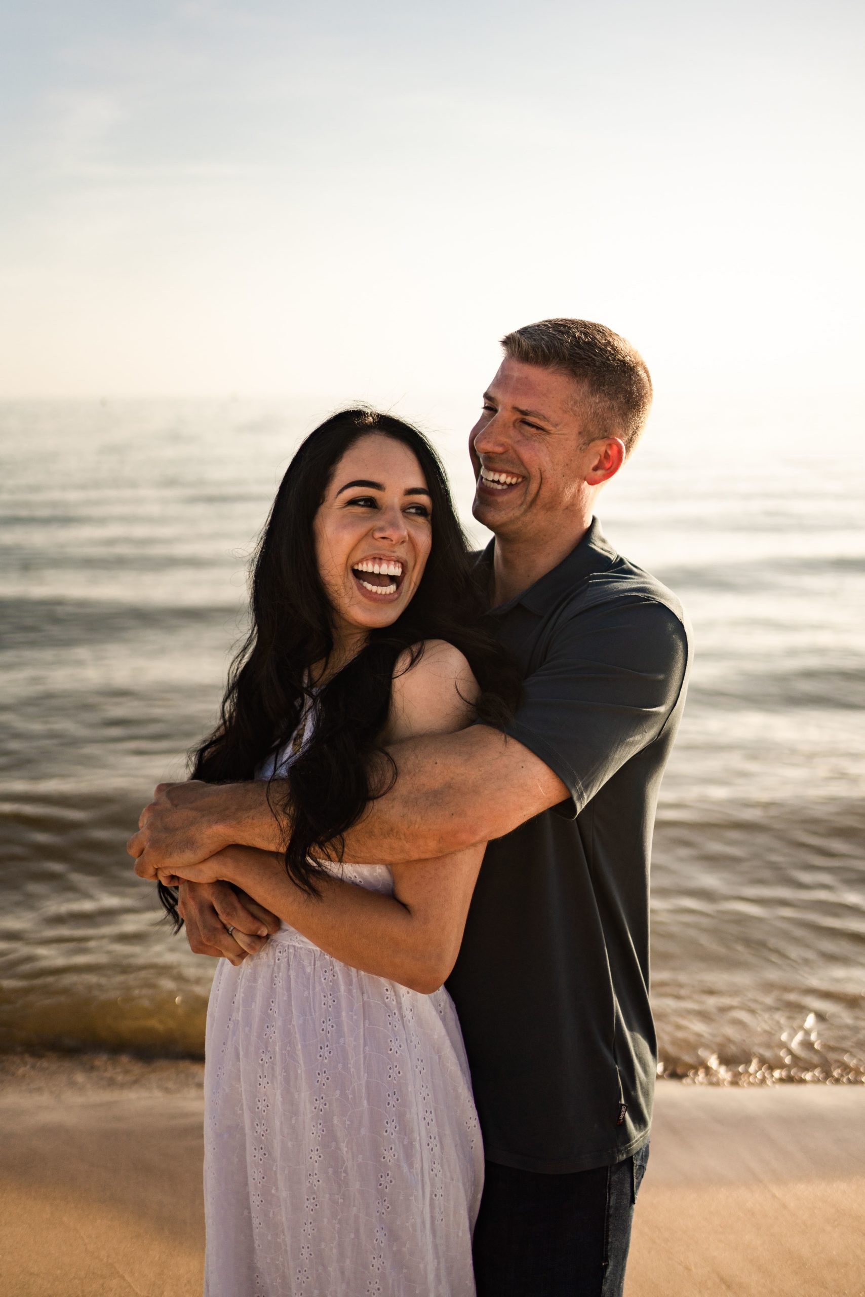 an engaged couple with wide smiles stand on the shore his arms wrapped around her during their engagement photoshoot beach