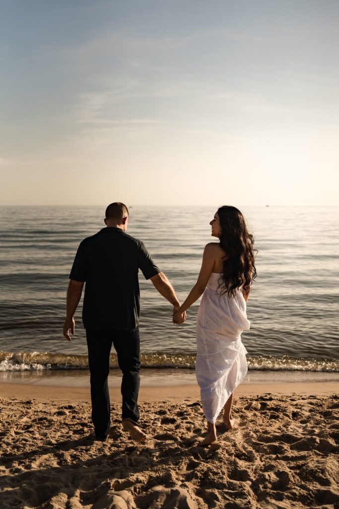an engaged couple hold hands as they walk towards the water during their engagement photoshoot beach